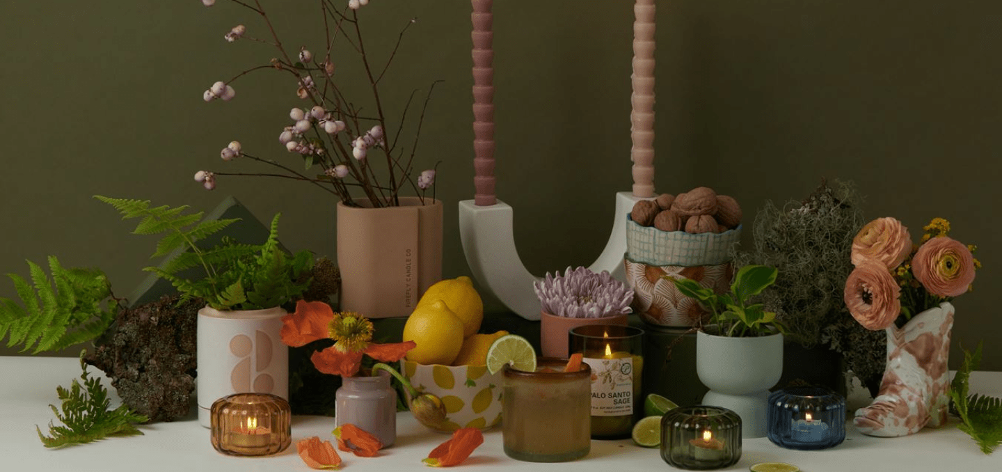 A collection of candles, vases with flowers, walnuts, lemons, and decorative objects arranged on a table against an olive-green background.