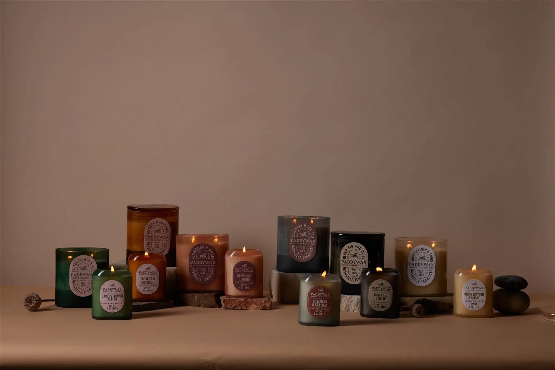 The Worn Leather + Smoke - Vista 12 oz Candle is displayed lit on a tan surface with a neutral background, accompanied by decorative stones and wooden sticks.