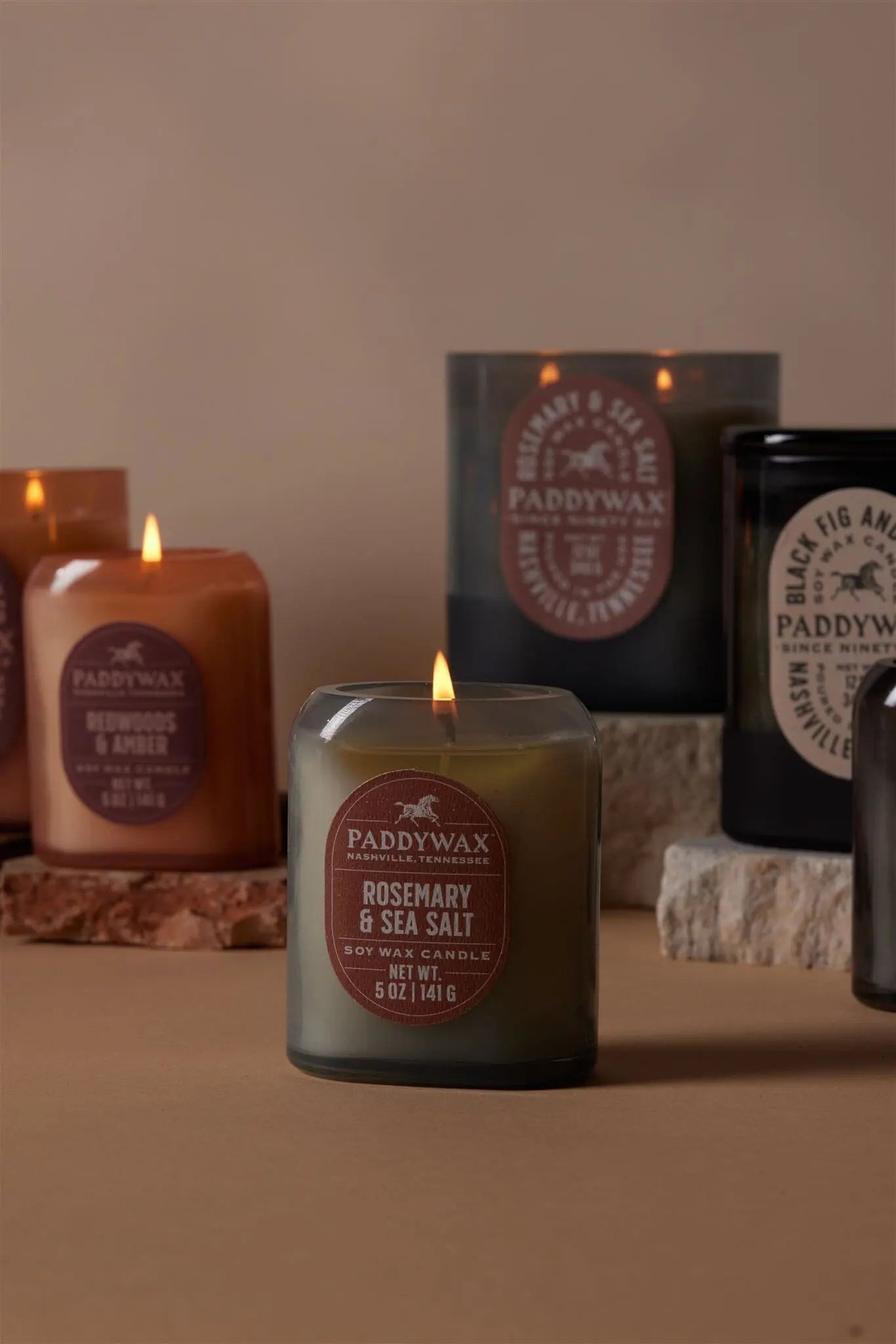 A lit Rosemary + Sea Salt - Vista 12 oz Candle from the vintage-inspired collection is displayed on a neutral surface with stone blocks.