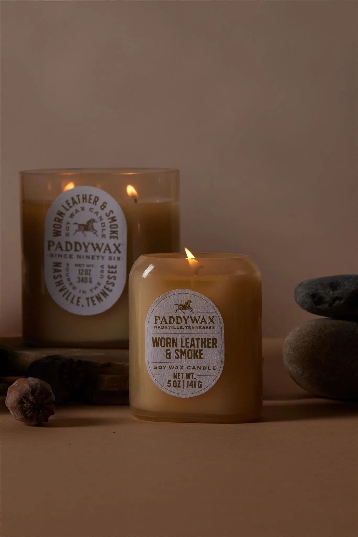 Two Worn Leather & Smoke - Vista 5 oz Candles are shown on a beige surface, one lit and one unlit, with gray stones and a dried seed pod—ideal for modern decor and adding a luxurious fragrance to your home.