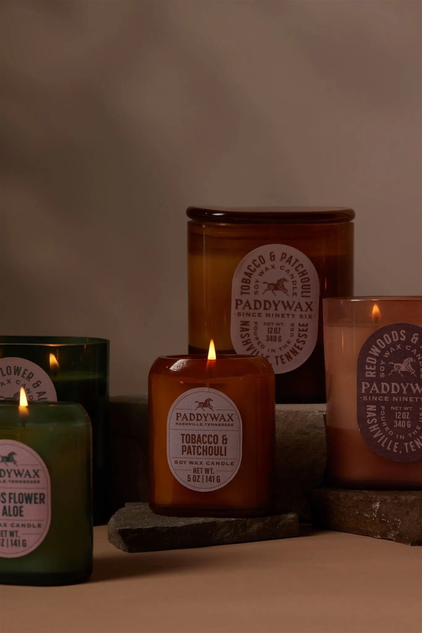 Five lit Tobacco + Patchouli - Vista 5 oz Candles by Paddywax in amber and green glass jars are arranged on stone slabs, bringing modern decor flair as their labels highlight the luxurious fragrance.