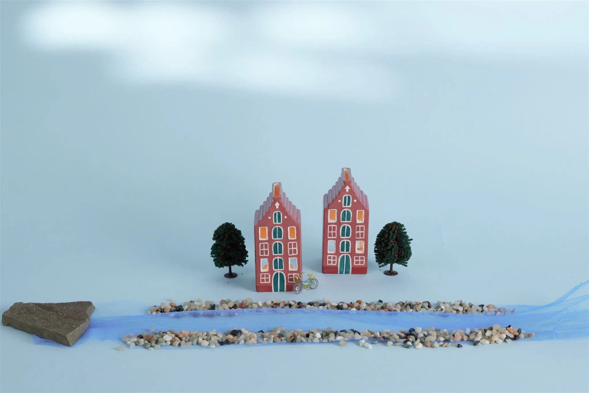 The No. 02 The Amsterdam House - Incense & Tea Light Holder is displayed with trees, pebbles, a blue fabric strip, and a rock on a light surface, capturing the charm of Amsterdam canal house decor.