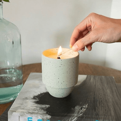 A hand lights a candle with a match. The candle is in a speckled ceramic holder and is placed on a book next to a glass bottle filled with water.