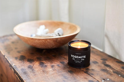 A Yosemite - Parks 6 oz Tin Candle glows on a rustic wooden surface beside a shallow wooden bowl filled with decorative items.