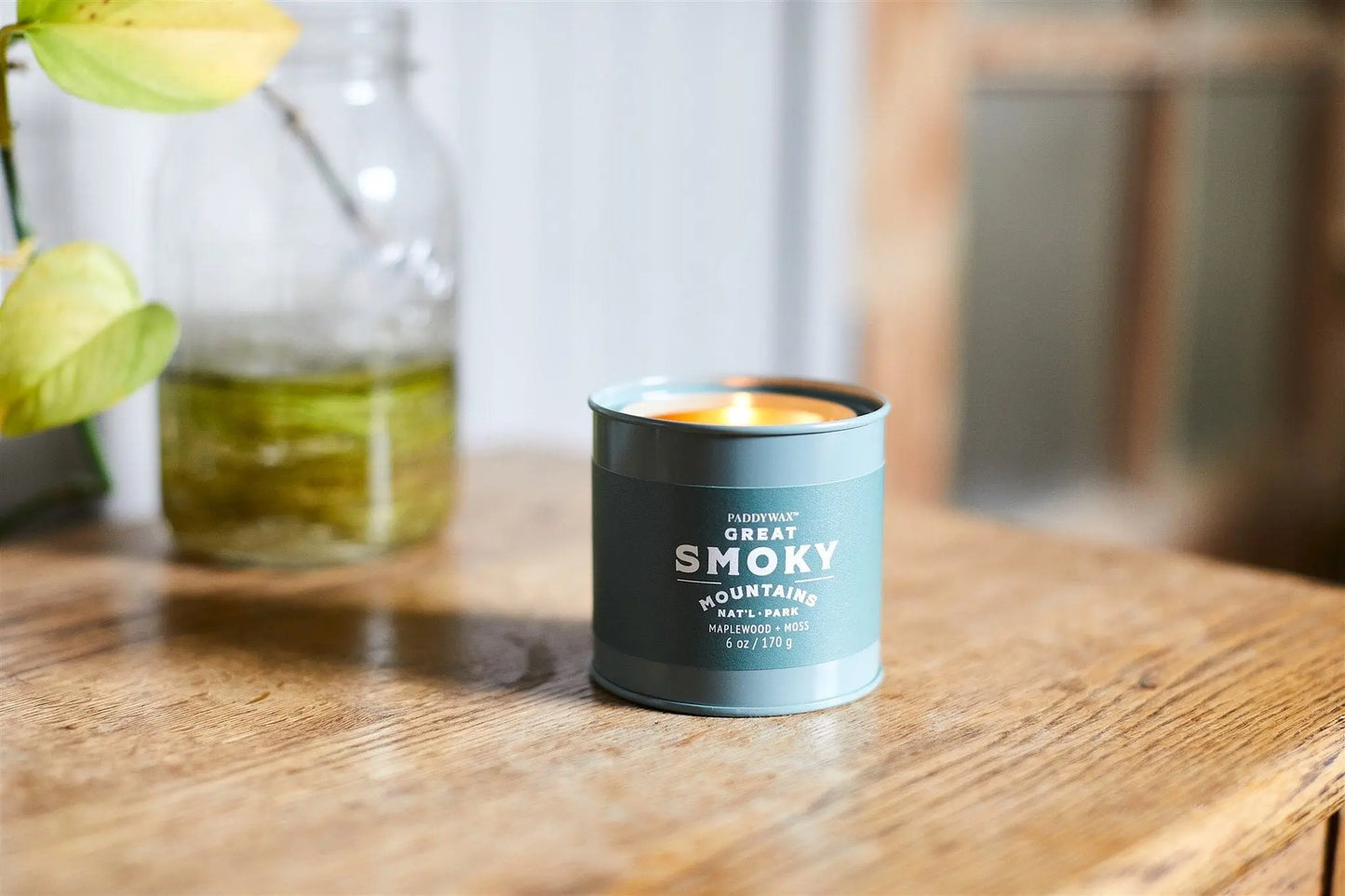 A Great Smoky Mountains - Parks 6 oz Tin Candle in blue sits on a wooden table beside a glass jar with green liquid and a plant leaf, making it an ideal gift for outdoor enthusiasts.