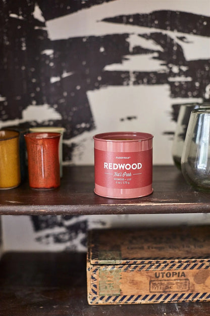 A Redwood - Parks 6 oz Tin Candle rests on a wooden shelf next to ceramic cups and glasses, above a UTOPIA box—an ideal outdoor gift for nature lovers.