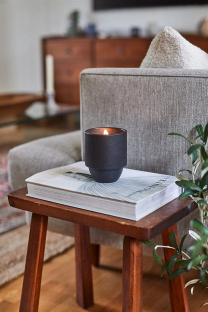 The Yosemite - Parks 11 oz Candle, featuring a woody Cottonwood and Oak scent and a crackling wood wick, burns atop a closed book on a wooden side table beside a gray armchair in a cozy living room.