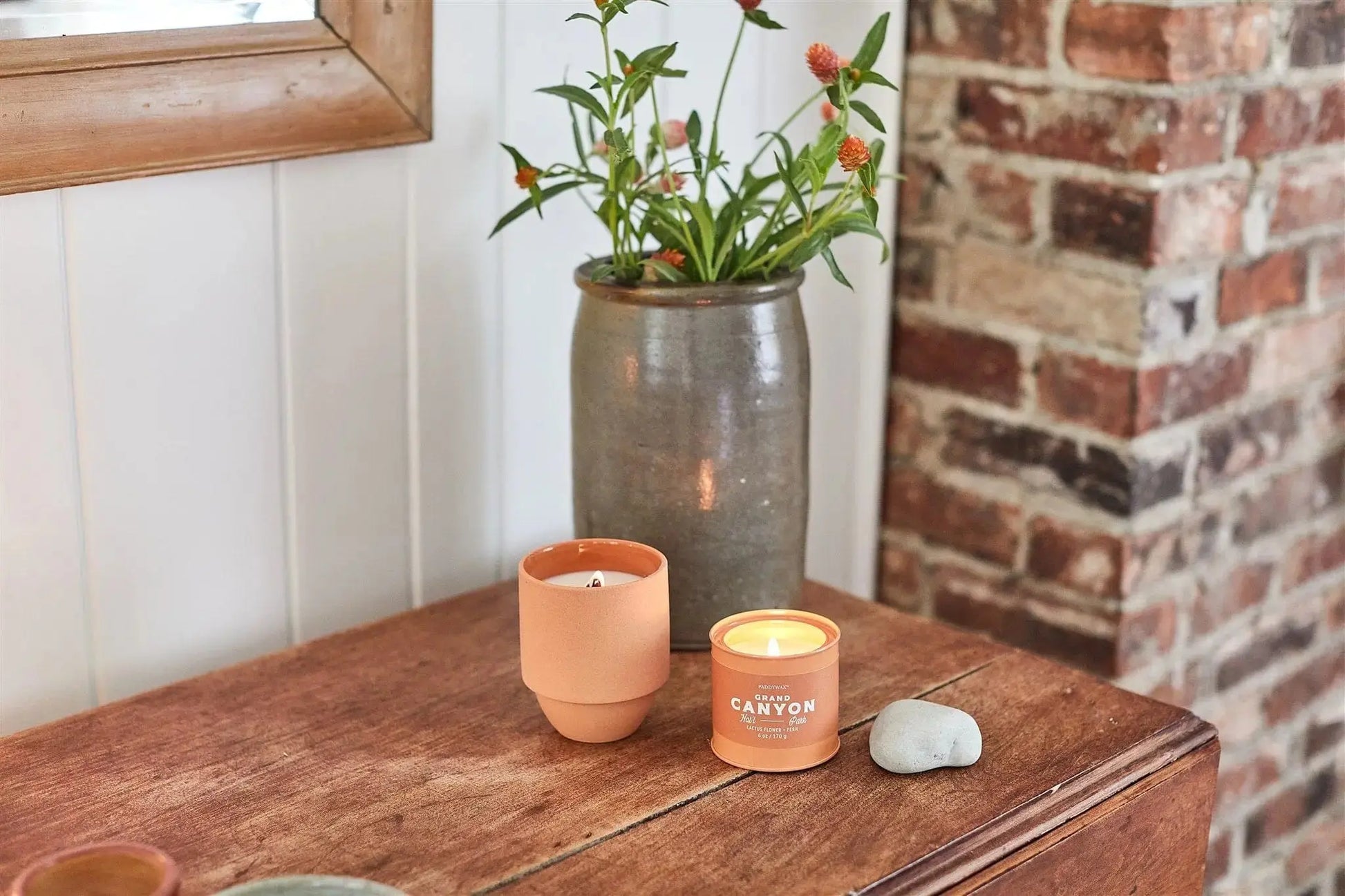A Grand Canyon - Parks 6 oz Tin Candle, a small planter, a smooth stone, and a vase with orange flowers sit on a wooden table by a brick wall—an inviting scene and an ideal gift for outdoor lovers from the Parks Collection.