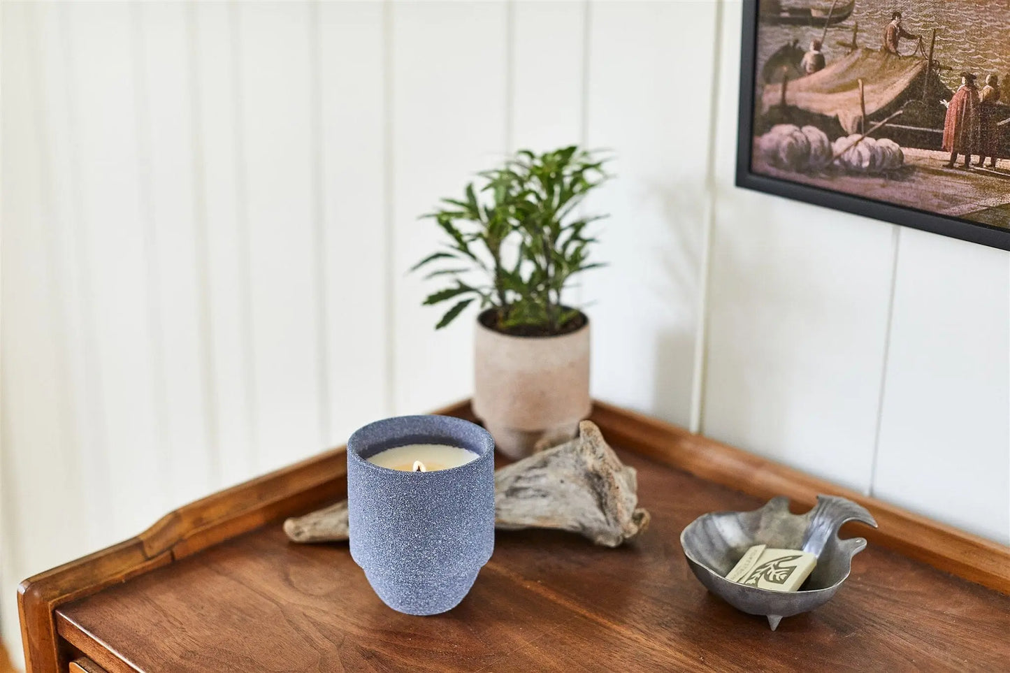 A small potted plant, driftwood, papers in a decorative dish, and the Acadia - Parks 11 oz Candle with a crackling wood wick are on a wooden table beside a framed picture on a white wall.