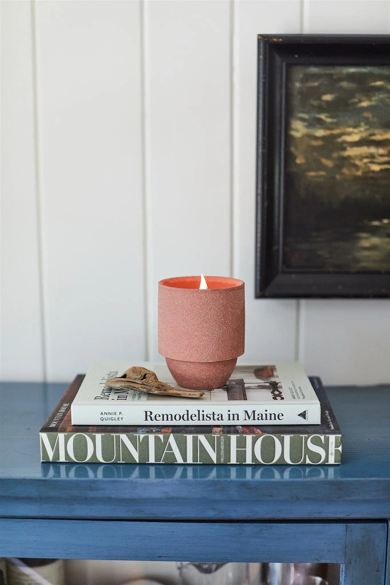 The Redwoods - Parks 11 oz Candle, featuring a crackling wood wick and pink wax, sits atop two books on a blue table. Its woody Redwood and Lily scent is inspired by Redwoods National Park. A framed painting appears in the background.