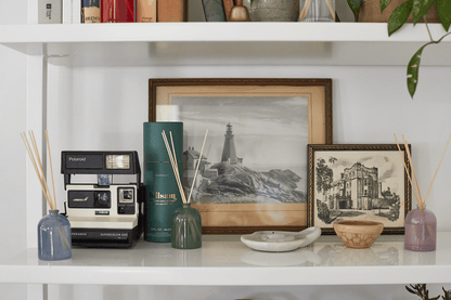 A shelf displays a Polaroid camera, framed lighthouse and building photos, books, a candle, bowl, ceramic dish, and the Balsam - 1.5 oz Diffuser—making it a perfect fragrance gift for any home.