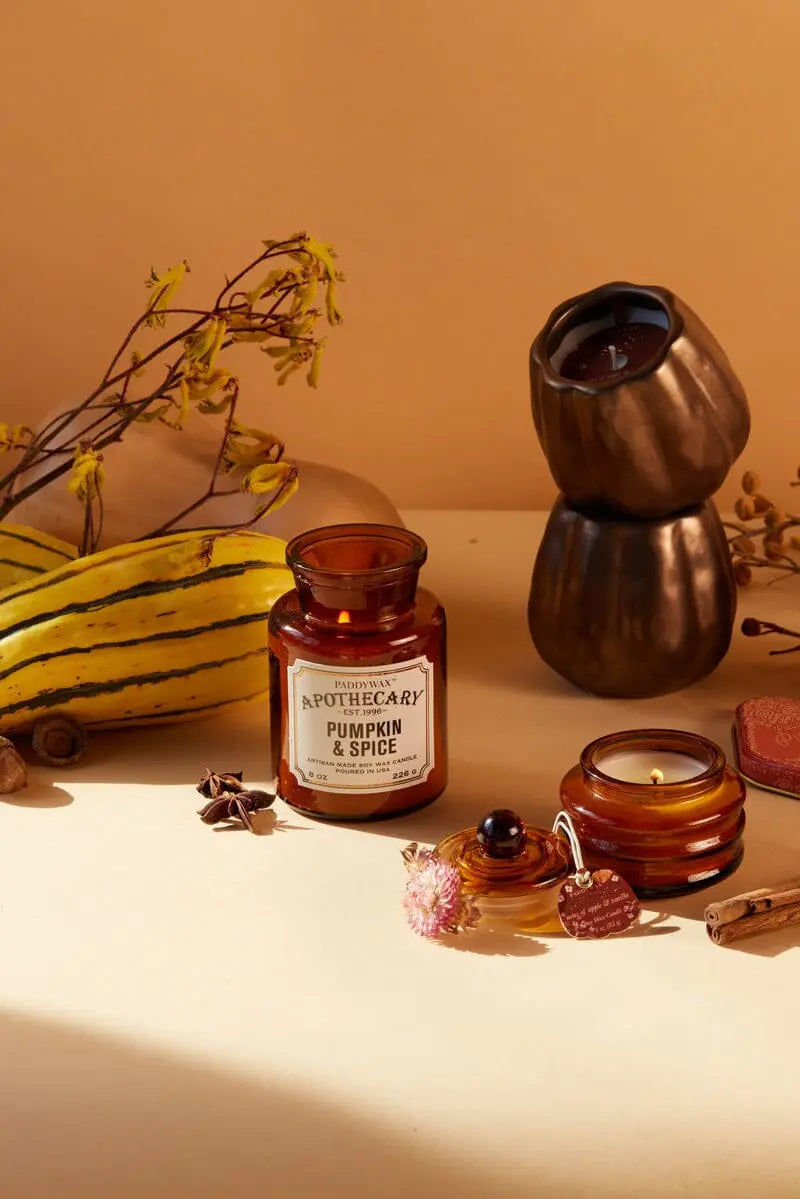 The Pumpkin + Spice - Apothecary 8 oz Candle rests in a brown glass jar on a beige surface, surrounded by gourds, a metal candle holder, dried flowers, and small decorative items.
