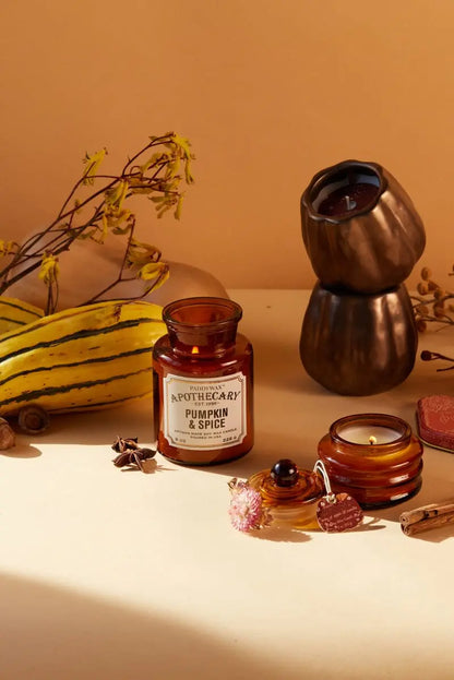The Pumpkin + Spice - Apothecary 8 oz Candle rests in a brown glass jar on a beige surface, surrounded by gourds, a metal candle holder, dried flowers, and small decorative items.