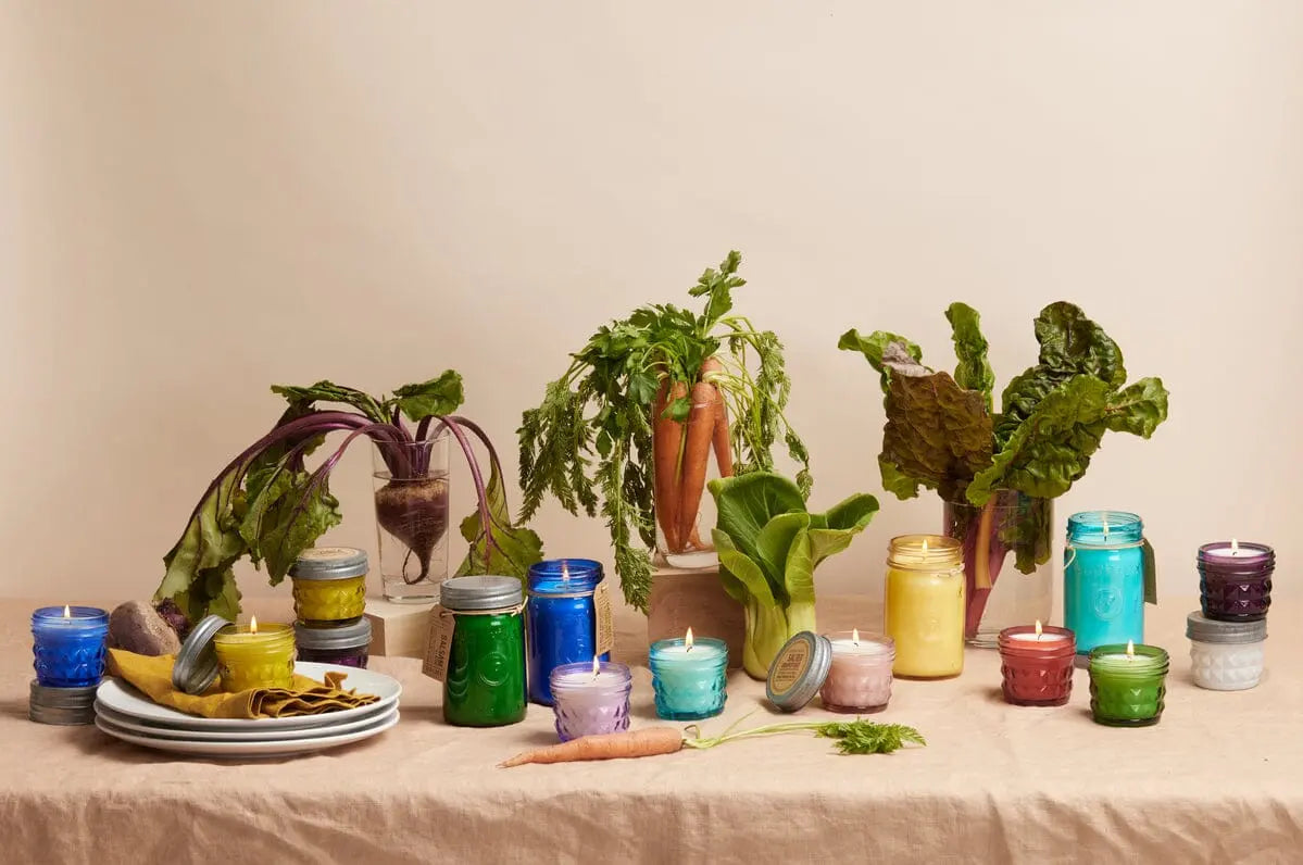 An Ocean Tide & Sea Salt - Relish 9.5 oz Candle in an upcycled glass jar, with fresh vegetables, is arranged on a beige tablecloth against a plain background.