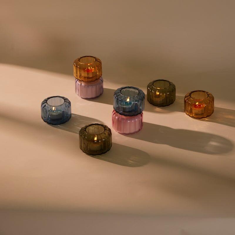 Several colored glass candle holders, some stacked, are placed on a beige surface with soft light casting long shadows.