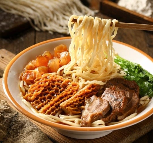 A bowl of noodle soup topped with beef tripe, braised beef, leafy greens, and chunks of meat. Chopsticks are lifting a portion of noodles above the bowl on a rustic wooden table.