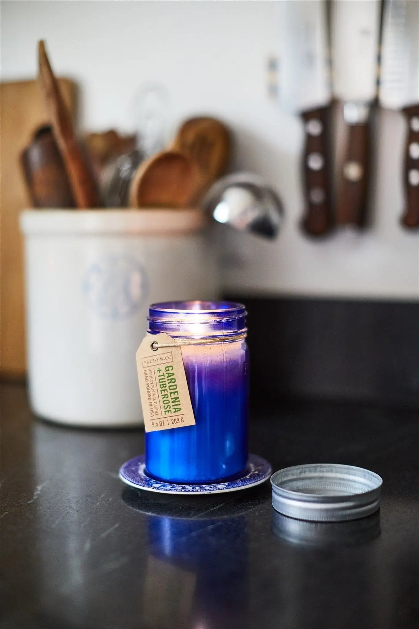 A lit Gardenia & Tuberose - Relish 9.5 oz Candle in a blue jar sits on a kitchen counter on a coaster, its metal lid nearby, with utensils and knives visible in the background.