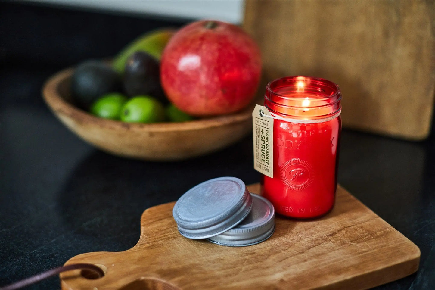 A Pomegranate & Spruce - Relish 9.5 oz Candle burns on a wooden cutting board with its metal lid nearby; in the background, a wooden bowl contains a pomegranate, limes, and avocados.