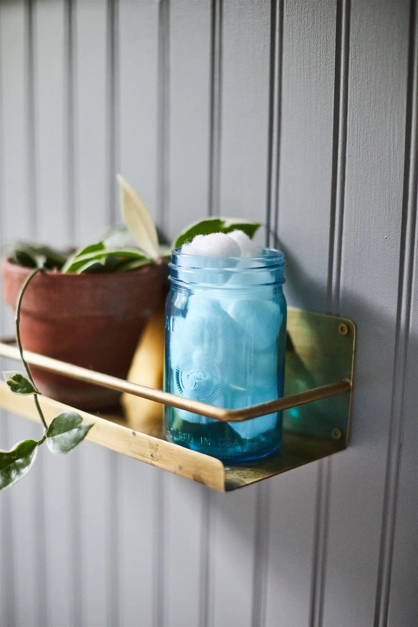 A blue Ocean Tide & Sea Salt - Relish 9.5 oz Candle sits on a brass shelf beside a potted plant and cotton balls against a gray paneled wall.