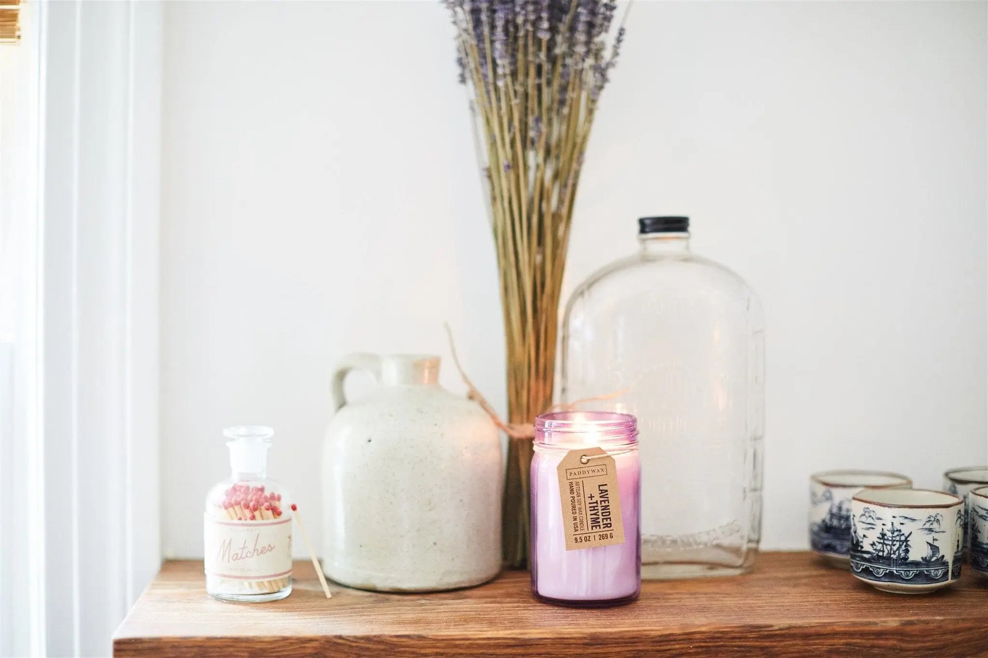 A wooden shelf holds a storage jar of matches, a ceramic jug, the Lavender + Thyme - Relish 9.5 oz Candle (lit), a glass bottle, blue and white cups, and dried lavender in a vase.