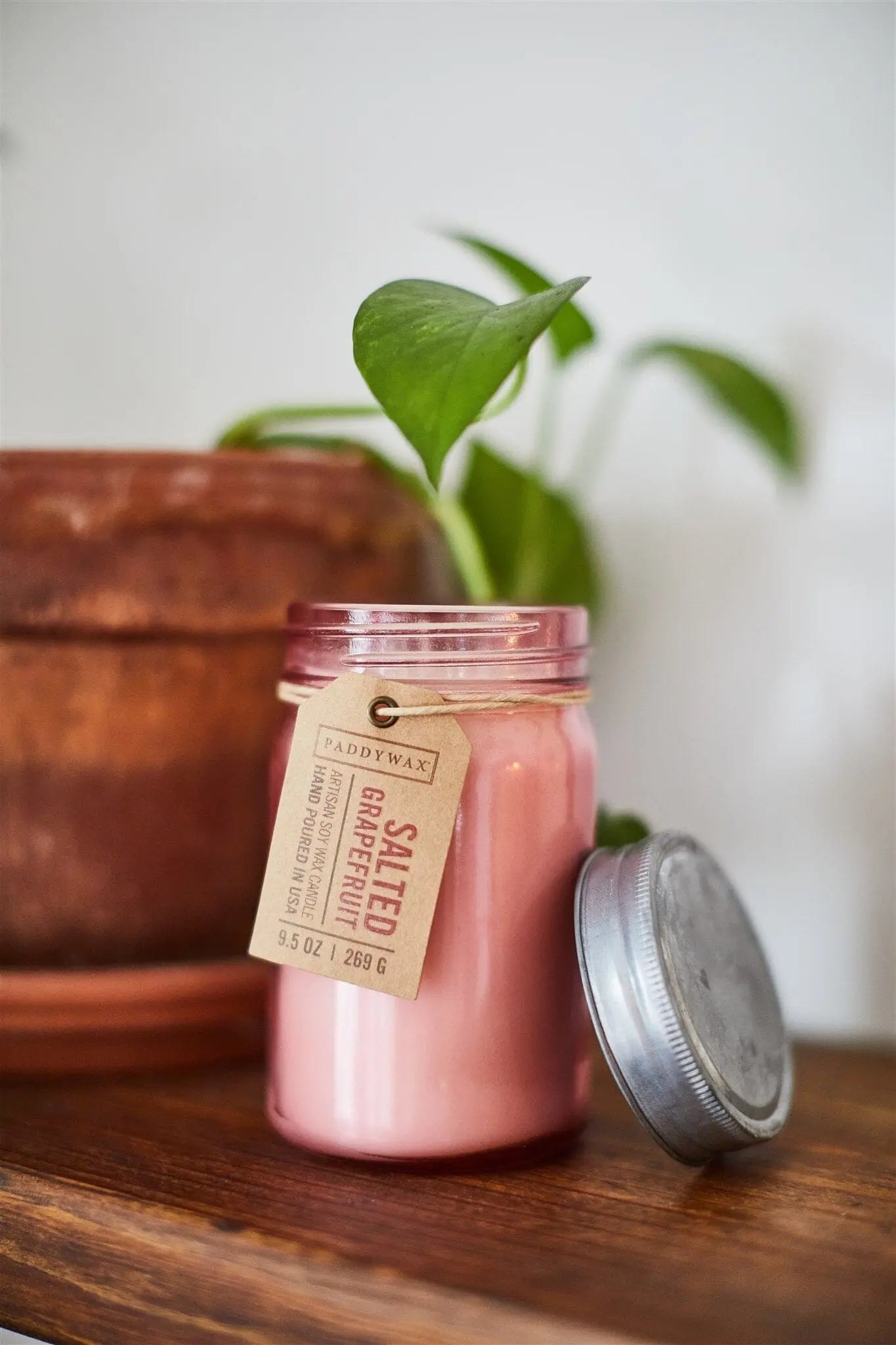 The Salted Grapefruit - Relish 9.5 oz Candle in pink is displayed with its lid off on a wooden surface beside a potted plant, offering a stylish and fragrant décor accent for any home.
