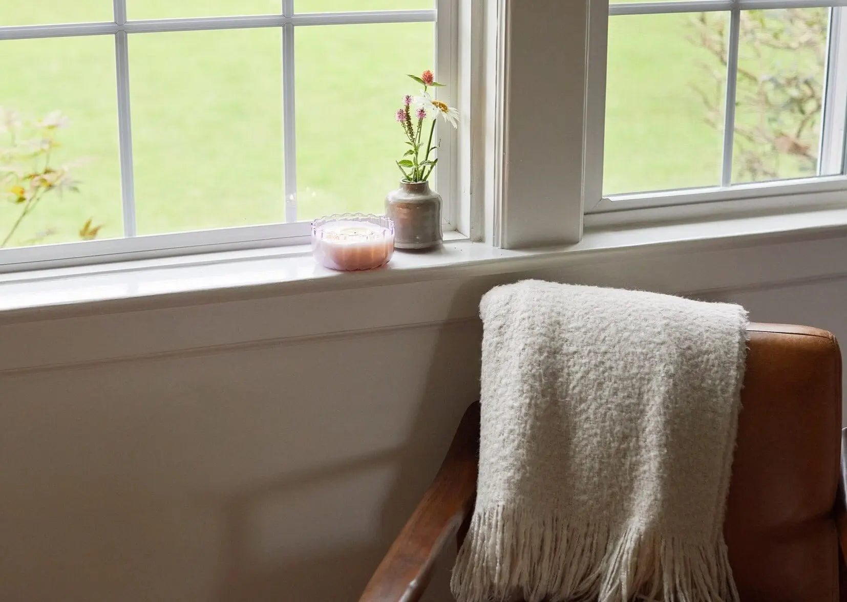 A white blanket drapes over a brown chair by the window, with a Salted Iris - 12 oz Candle and a small vase of flowers placed on the windowsill.