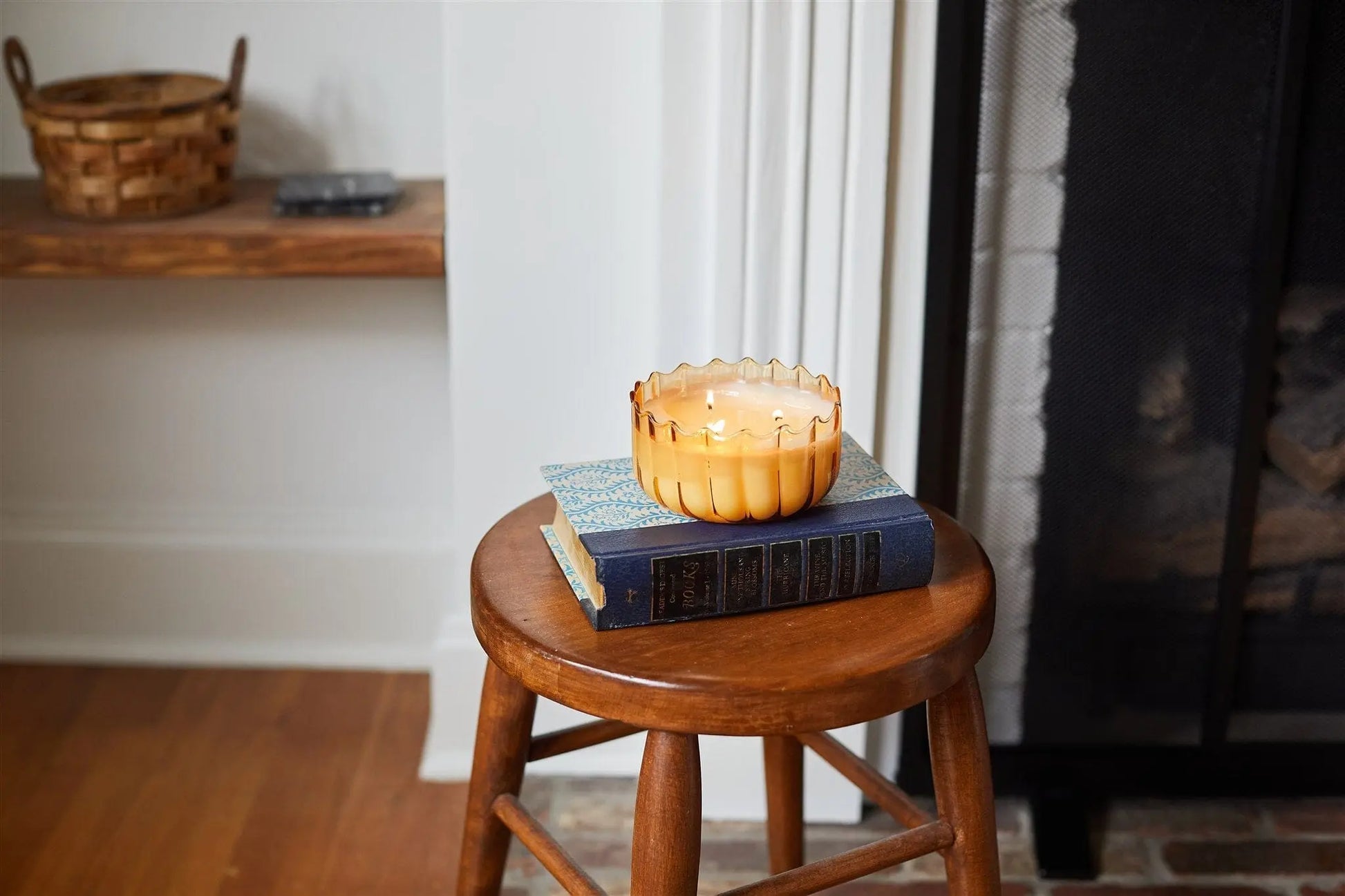 The Golden Ember - Ripple 12 oz Candle in a ribbed glass holder sits on two books atop a wooden stool, bringing vintage charm as unique table decor near the fireplace.