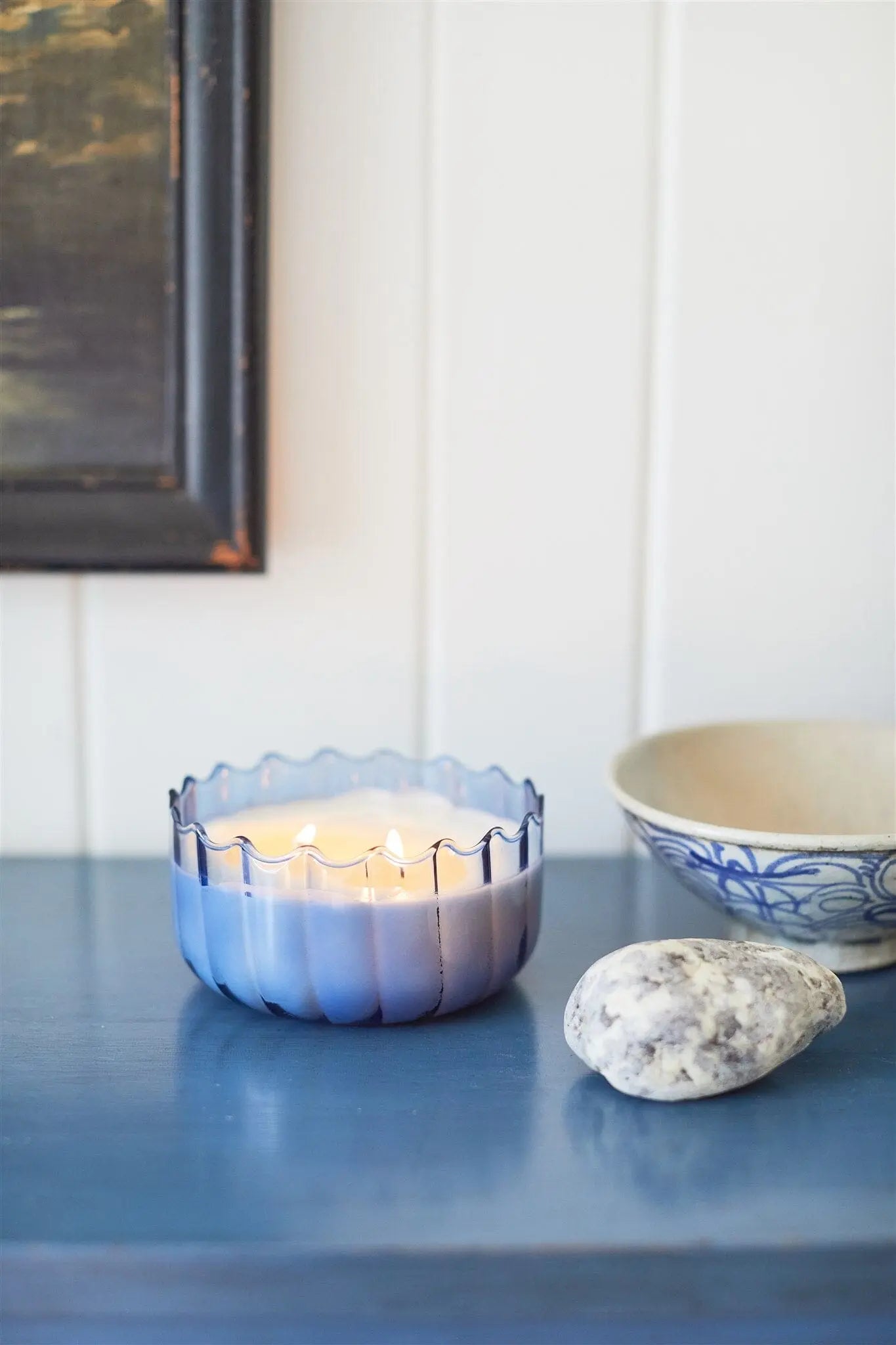 The Peppered Indigo - Ripple 12 oz Candle in a ribbed glass holder rests on a blue surface near a gray stone and a white bowl decorated with blue patterns.