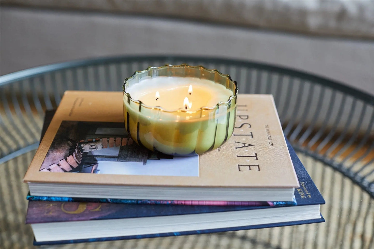 A lit Secret Garden - Ripple 12 oz Candle in a green glass container rests atop two stacked books on a round glass table.