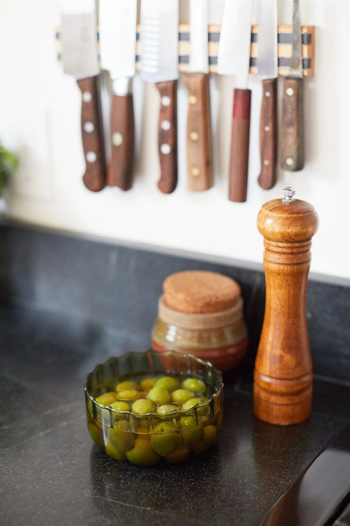 A bowl of green olives sits on a black countertop near a wooden pepper grinder, a jar with a cork lid, and the Secret Garden - Ripple 12 oz Candle beside a magnetic knife strip on the wall.