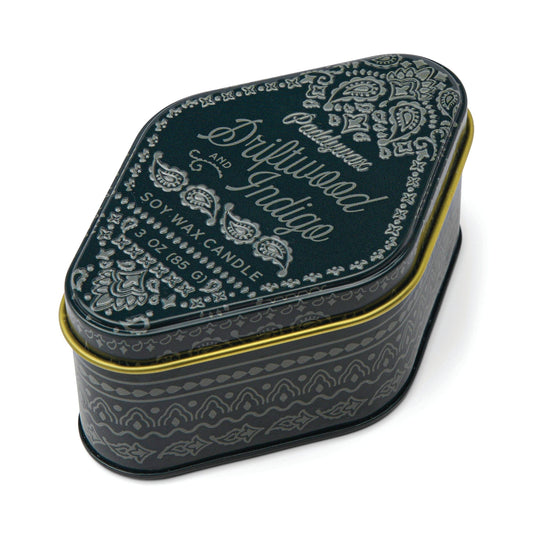 A dark, teardrop-shaped tin with ornate white paisley designs and a gold rim, the Driftwood Indigo - Station Tin 3.5 oz features “Driftwood and Indigo Soy Wax Candle” on its lid. Perfectly sized for travel, this candle is inspired by National Parks scents and fills any space with a woody aroma reminiscent of tranquil forest trails—making it an ideal choice among travel-sized tin candles.