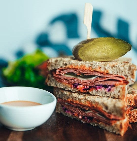 A stacked sandwich with sliced meat, cheese, and vegetables on whole grain bread, topped with a pickle and held by a skewer, sits beside a small bowl of sauce and leafy greens on a wooden surface.