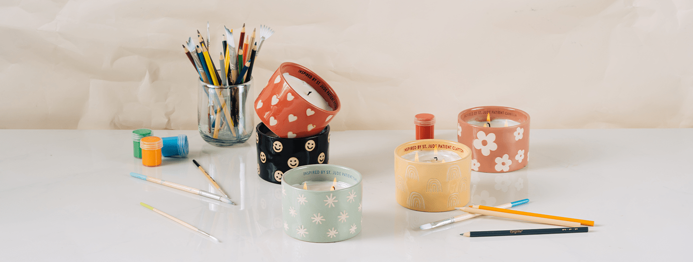 A variety of colorful candles in decorated jars sit on a white surface, surrounded by colored pencils, pens, and paintbrushes in a glass jar. The scene is creative and cheerful.