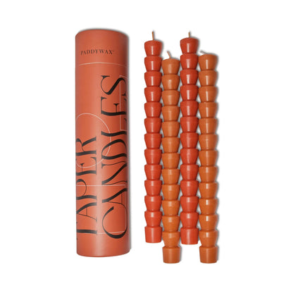 A tall cylindrical box labeled Red & Terracotta Tube Tapers - Set of 4 stands beside four stacks of short, orange-red contemporary tapers—ideal for adding vibrant color to modern table settings on a white background.