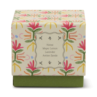 The Bronzed Gardenia - Tulip 5.5 oz Candle comes in a decorative box with a vibrant floral design and green base with white dots. Label notes: Meyer Lemon, Lavender, Amber Sands—perfect for floral home decor.
