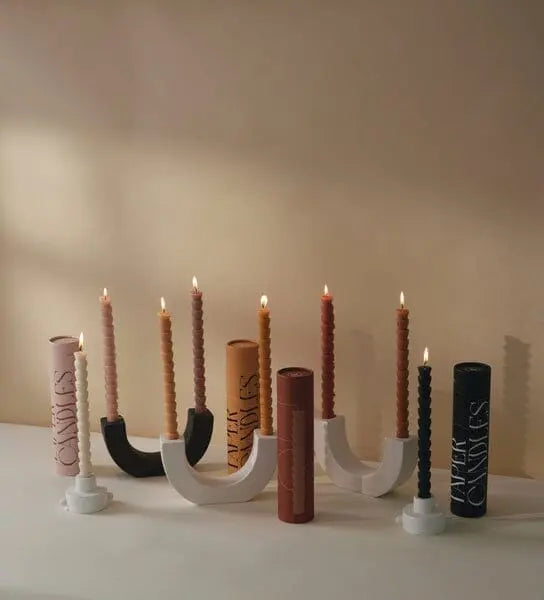 A row of lit Red & Terracotta Tube Tapers sits in modern U-shaped and circular candle holders, with matching taper boxes displayed behind them on a light-colored surface.