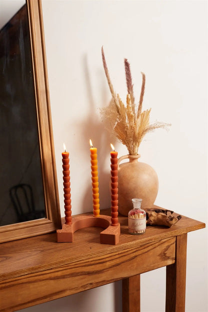 A wooden table with a mirror, a set of four red & terracotta tube tapers in a curved holder, a vase with dried grasses, a small decorative bottle, and a wooden cat figurine on top.