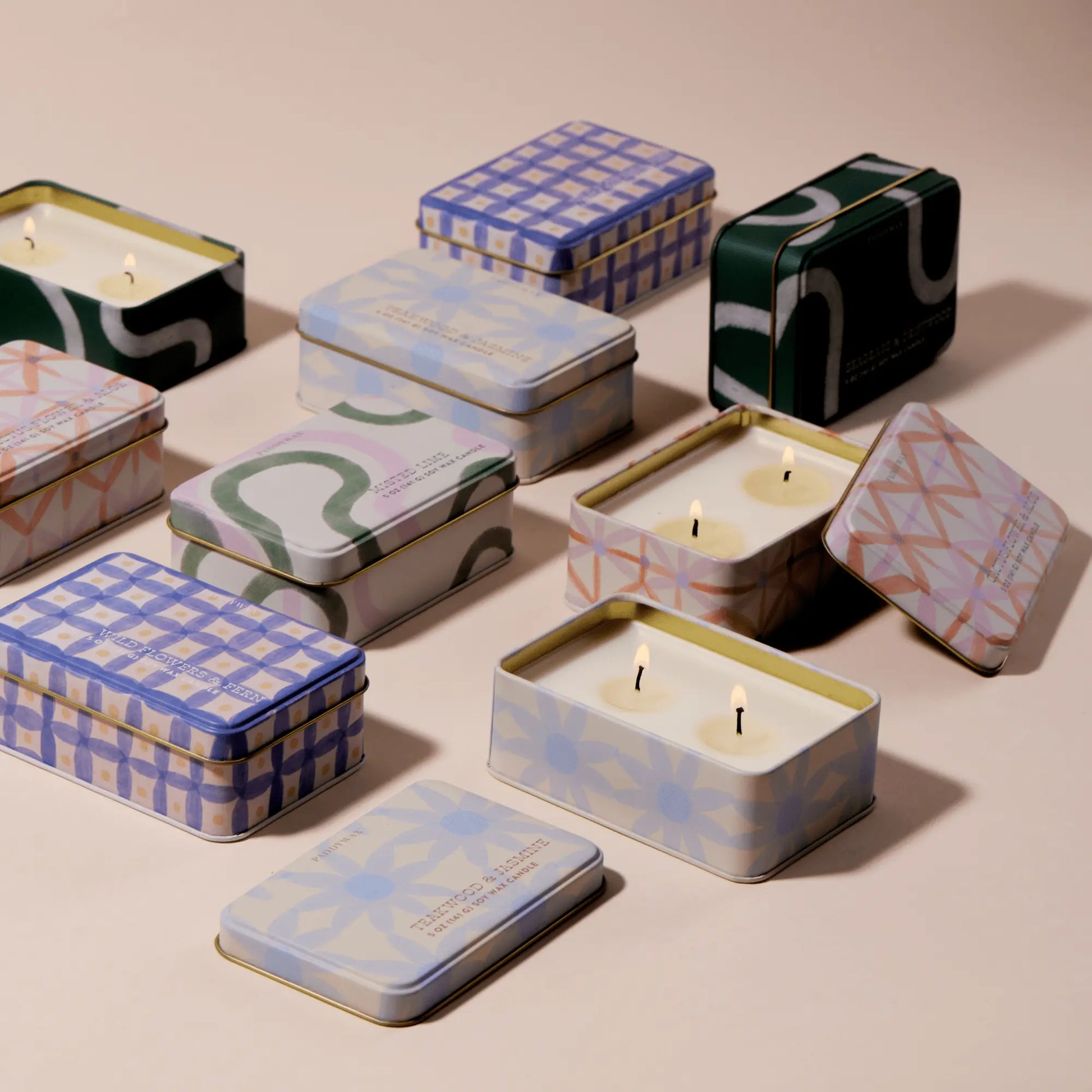 Rectangular tin candles in pastel colors and patterns sit on a beige surface, some lidded, others revealing wax. The Seagrass & Driftwood - 5 oz Tin Candle lends a fresh, coastal vibe to the collection.