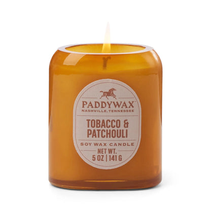A lit Tobacco + Patchouli - Vista 5 oz Candle adds modern home decor style and a luxurious scent against a clean white background.