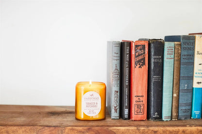 The lit Tobacco + Patchouli - Vista 5 oz Candle adds modern charm to a wooden surface, filling the air with a luxurious scent beside upright hardcover books.