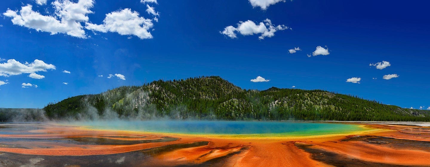 A vibrant hot spring with orange, yellow, and blue colors steams under a bright blue sky, surrounded by forested hills and scattered clouds.
