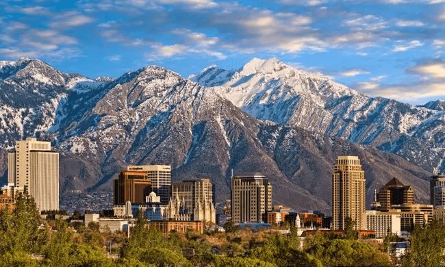 Downtown city skyline with tall buildings in front of rugged, snow-capped mountains under a partly cloudy sky. Green trees are visible in the foreground.