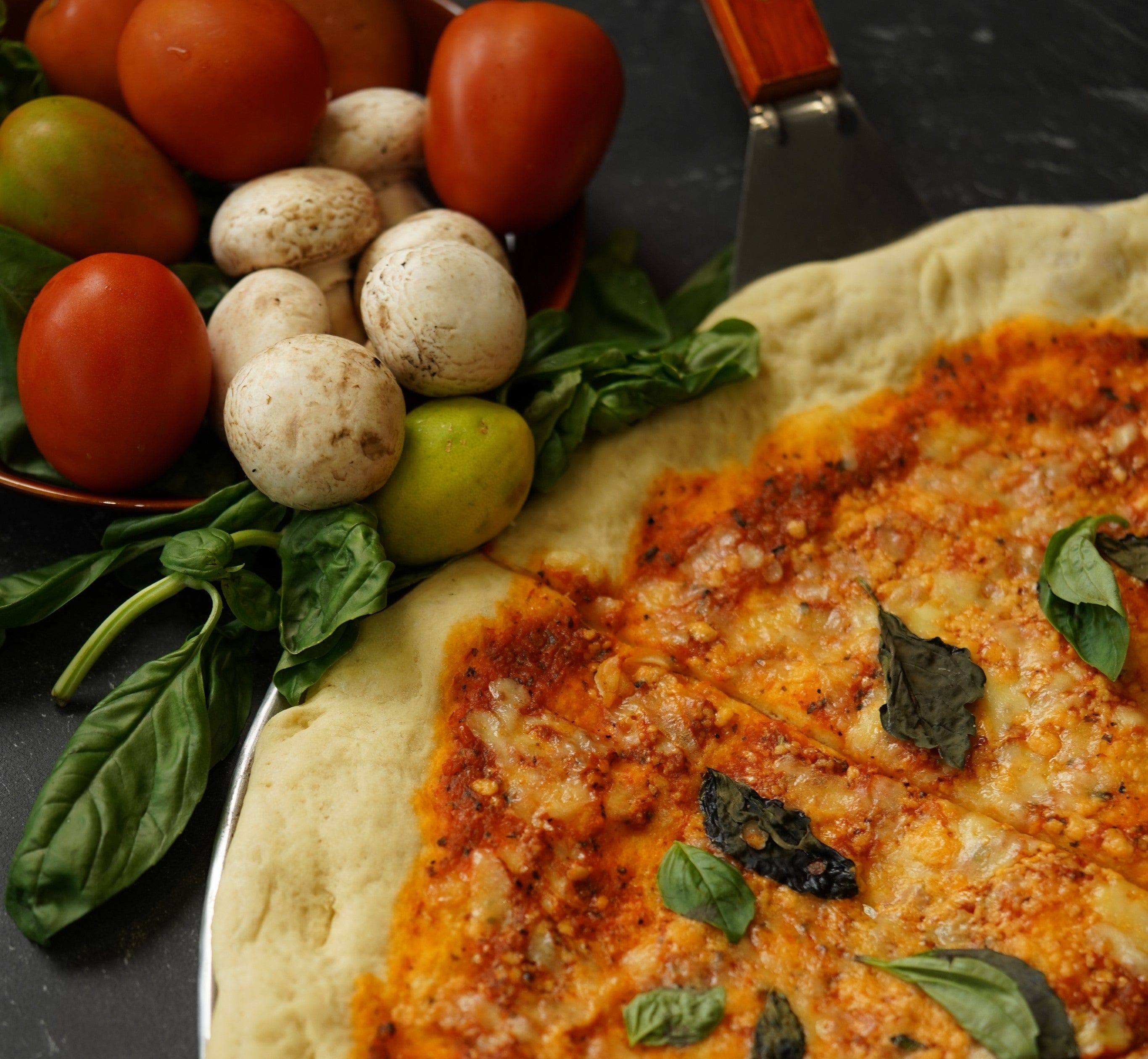 A close-up of a cheese pizza garnished with basil leaves, next to fresh tomatoes, white mushrooms, and basil on a dark surface. A pizza cutter is also partially visible.