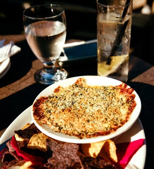 A bowl of baked dip with a crispy topping sits on a plate with assorted tortilla chips. In the background, there is a glass of water, a glass of iced drink with lemon, and a phone on the table.