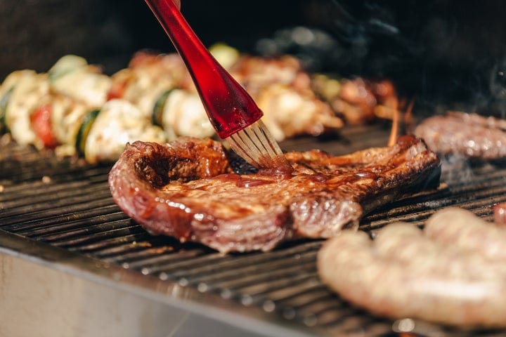 A close-up of a barbecue grill with steaks, sausages, and vegetable skewers. A red brush is being used to apply sauce to a steak, and smoke is rising from the grill.