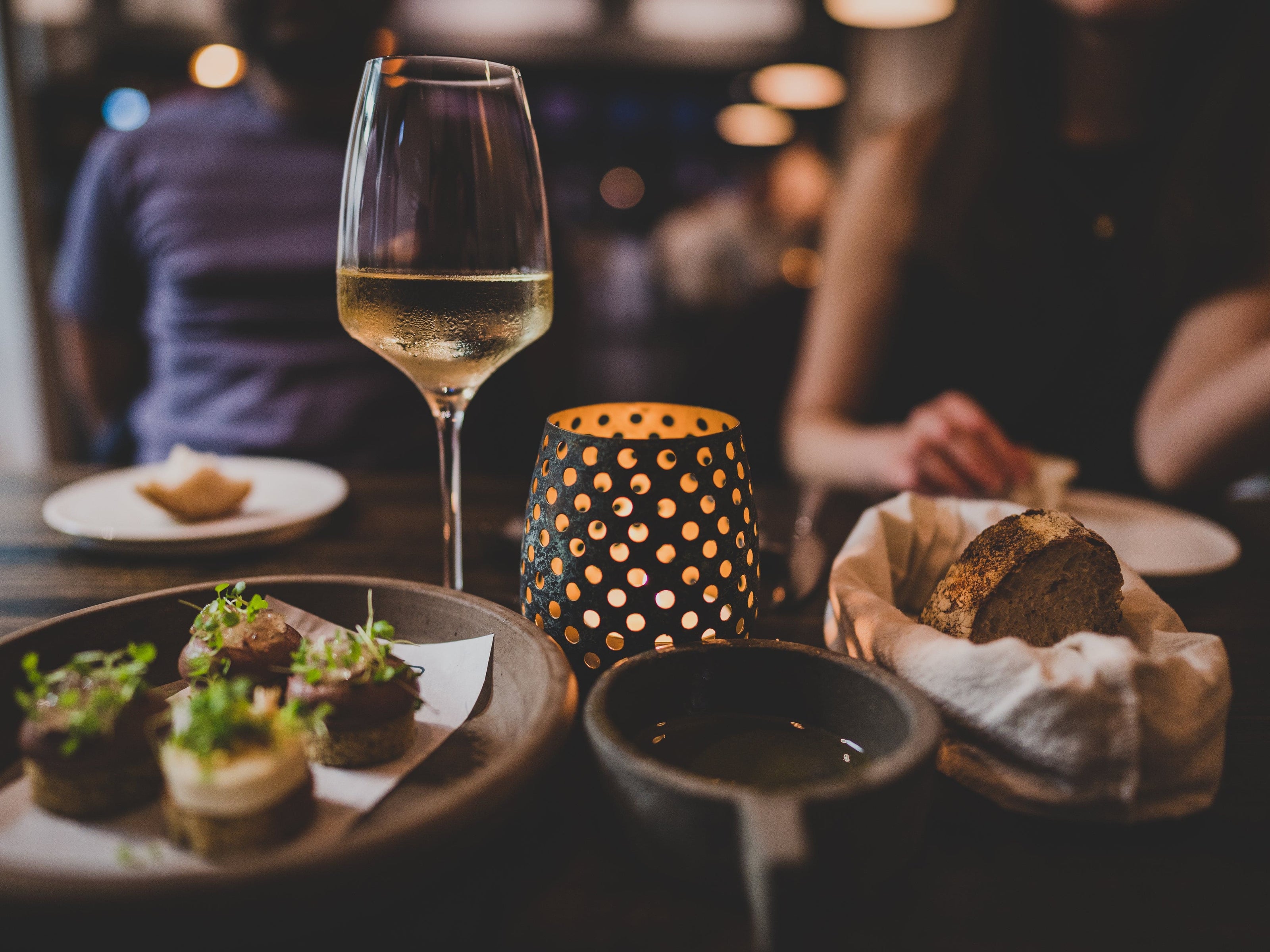A close-up of a dinner table set with a glass of white wine, a lit candle in a perforated holder, assorted appetizers, a bowl of olive oil, and rustic bread. People are sitting in the dimly lit background.