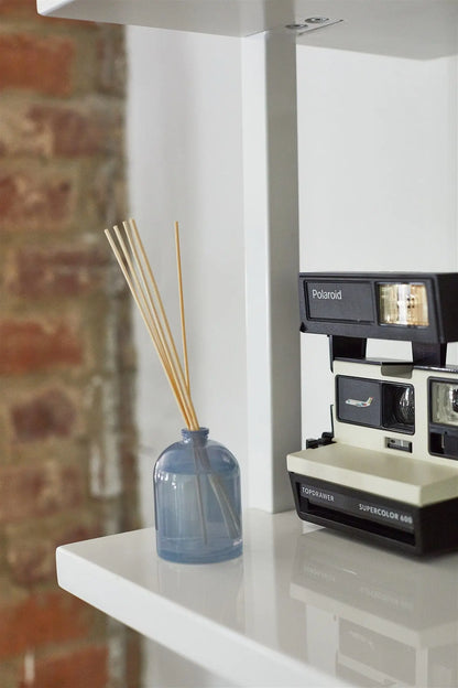 A Wild Fig - 1.5 oz Diffuser and a vintage Polaroid Supercolor 669 camera rest on a white shelf, creating a charming fragrance gift idea with a brick wall backdrop.