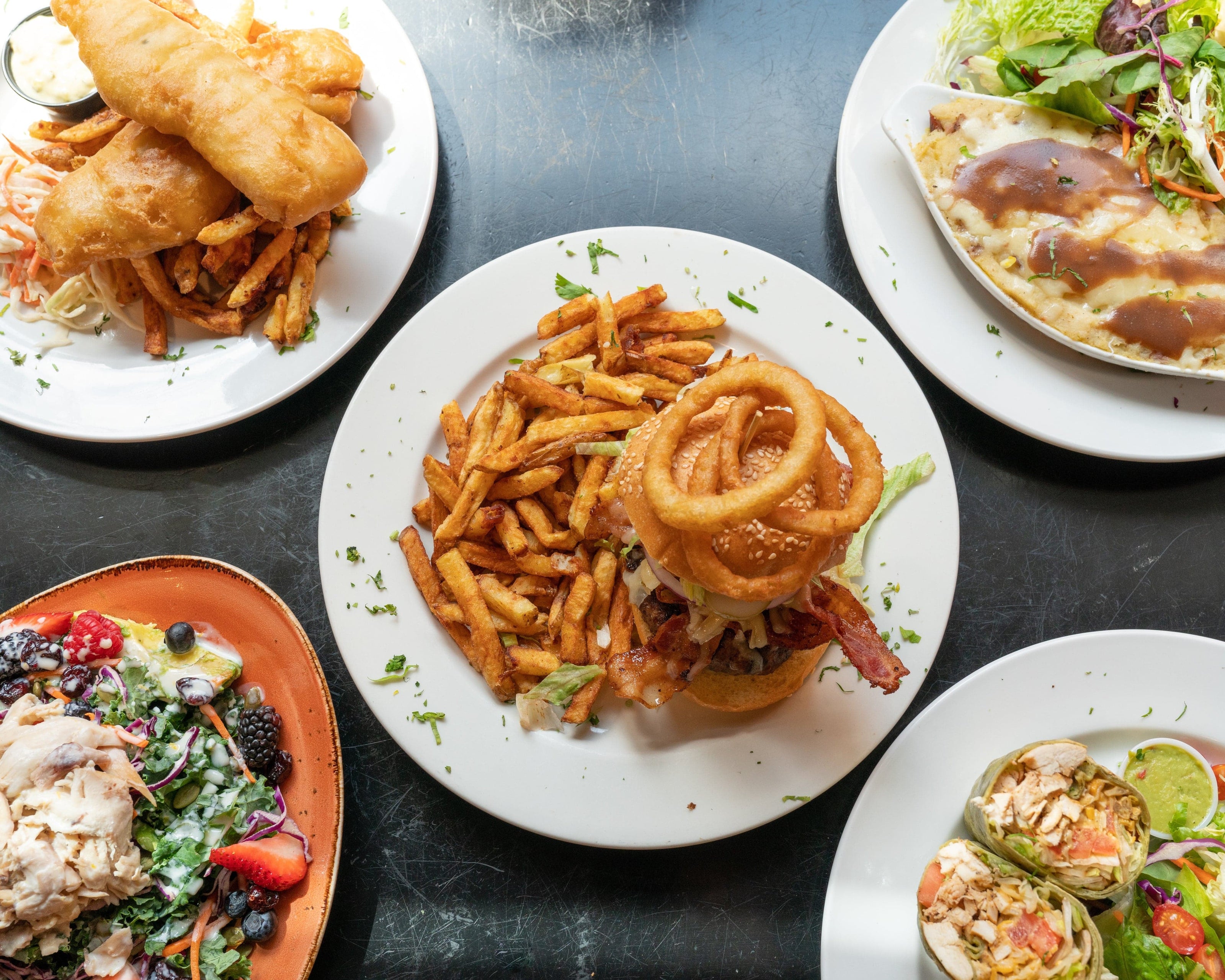 Overhead view of assorted dishes on a dark table, including a burger with fries and onion rings, fish and chips, a salad with berries, a wrap, and a plate with gravy-covered meat and greens.