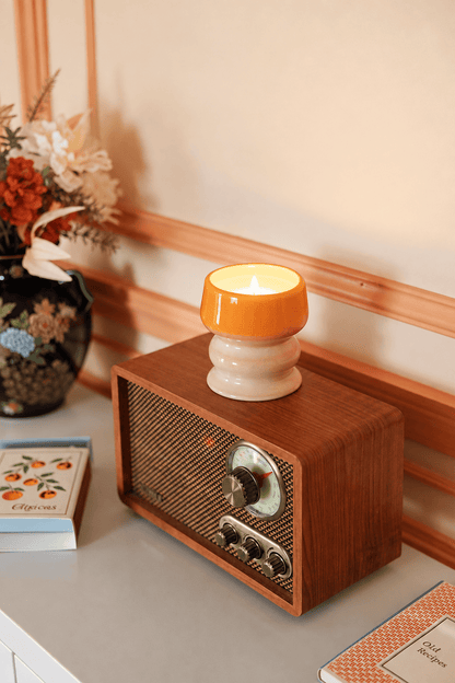 A vintage radio sits on a table beside a hand-glazed ceramic vessel and two closed notebooks with decorative covers, while the air is filled with the tropical scent of the lit Infrared Mango - Melt 8.5 oz Candle.