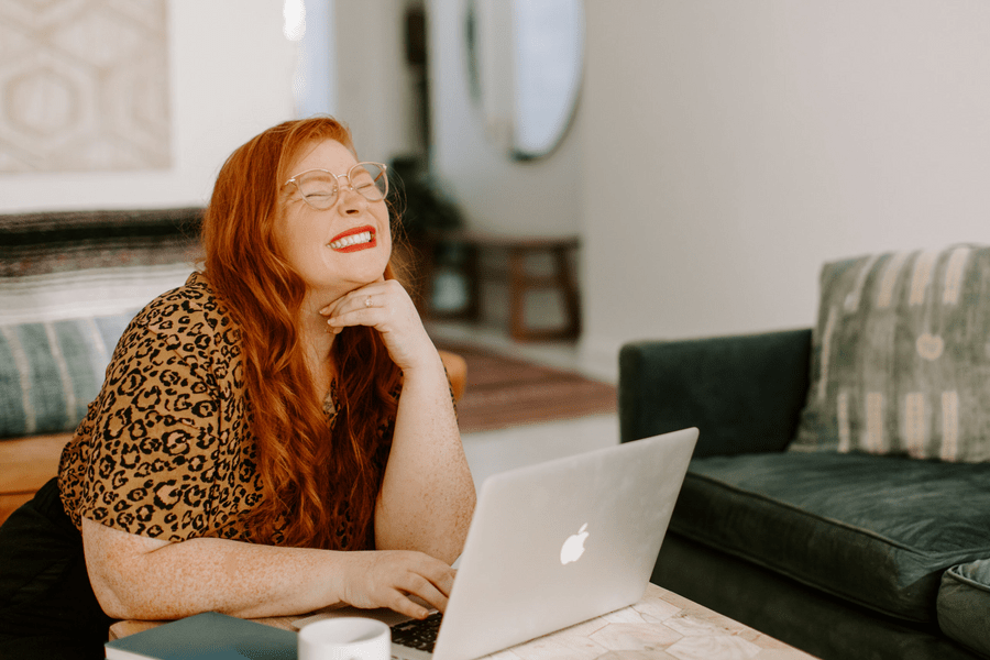 A woman with long red hair and glasses sits at a table, smiling with her eyes closed and hand under her chin, in front of an open laptop in a cozy living room.